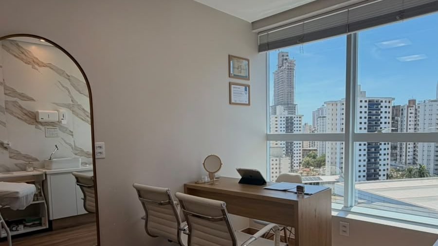 Sala de atendimento com espelho em arco, mesa de consulta e janela ampla com vista para os prédios do Jardim Goiás